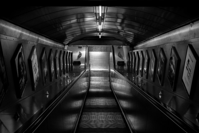 Monochrome photo looking down an single escalator, shiny metal stairs between shiny metal balustrade panels under a curved shiny metal roof, all pointing down to a shiny metal plate at the bottom. A row of posters runs down each side.