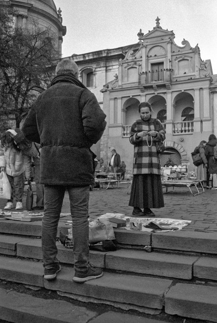 In this black-and-white street scene from a flea market, a man stands on the stone steps with his back to the camera, quietly observing a small spread of items laid out at his feet — books, trinkets, and bits of vintage clutter. Across from him, a woman in a long checkered coat and skirt carefully examines an object in her hands, absorbed in the moment.

Behind them rises an old, ornate building with arched balconies and decorative facades, adding a sense of history and stillness to the scene. Around the edges, other vendors and visitors move about, bending over tables and crates, creating a subtle bustle that contrasts with the calm interaction in the foreground. The grainy texture of the photo enhances the feeling of a timeless, everyday ritual taking place in an old European square.