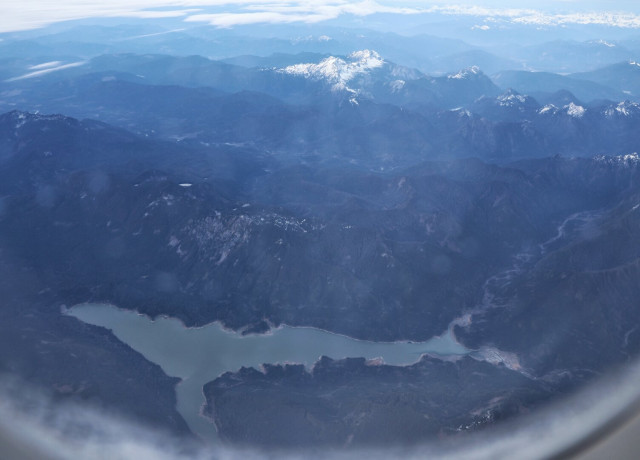 A stunning aerial view of a mountainous landscape, photographed from an aircraft window. Dominating the scene is a winding lake, its shape remarkably resembling that of a humpback whale from above. The lake’s calm, light blue waters contrast with the dense, shadowy valleys and forested hills that surround it. In the distance, towering peaks—some dusted with snow—rise against a hazy horizon, where layers of mist and clouds add a sense of depth and serenity to the scene.