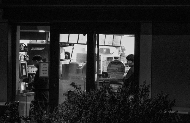 Une photographie de rue nocturne, en noir et blanc et au format paysage. Par une baie vitrée éclairée, on voit l'intérieur d'un lieu de restauration rapide. où de grands faitout exhalent un peu de vapeur. Sur la gauche, un homme concentré sur sa tâche se sert d'un distributeur de boisson et sur la droite de de cette fenêtre un homme désoeuvré est appuyé contre un mur. L'ensemble dégage une impression de tristesse anonyme.
