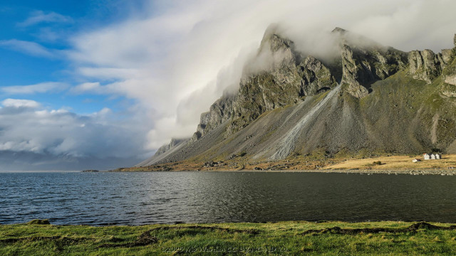A colour photo of a landscape with a body of water in the foreground. A large mountain is on the opposite side, a cap cloud hugging its peaks. The sky beyond this cloud is clear cyan.