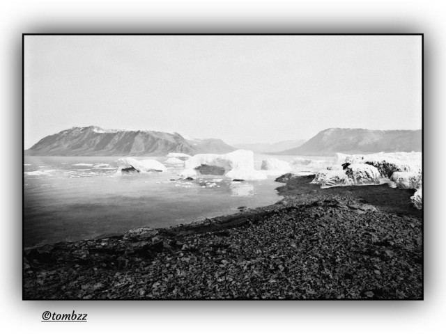 A black and white photo of Greenland’s southern coast. In the foreground, there’s a rough, rocky shoreline with cracks and uneven textures. Just beyond it, calm icy water stretches out, dotted with big chunks of floating ice...some look like broken-off glacier pieces, others like mini icebergs. In the distance, snow-covered mountains rise up, their edges softened by mist or fading light. The whole scene feels cold, quiet, and remote. The soft vignette adds depth and a sense of isolation. It’s all about texture, contrast, and the stark beauty of this frozen landscape.
