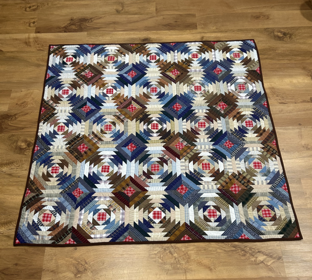 A pineapple style quilt with alternating diagonals of browns and blues in shirting patterns, mostly stripes and plaids. The middle sections are various white and cream shirtings again with stripes and crosshatching. The center squares and corners for each block are bright red with white cross-hatching. The pineapple effect made up of jagged lines hwich can be seen as forming pineapples but also interlocking circles.