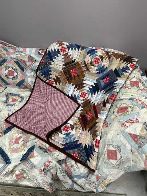 The quilt is lying, folded, on top of a well-worn pineapple quilt. Its corner is turned back to reveal the pink check on the back. The worn pineapple quilt looks almost like a shadow of the new one.