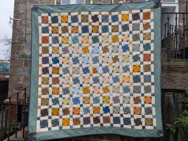 Patchwork quilt made up of an 11*11 grid of blocks with a windmill motif in colours mainly blues, yellows and mustards, the blocks on a white background with smaller coloured squares to corners. Centre block has a chicken. Tones of the blocks are darker to the edge, lighter to the quilt centre. All finished with a light blue border. Quilted in the ditch on the diagonal, picking up the windmills.