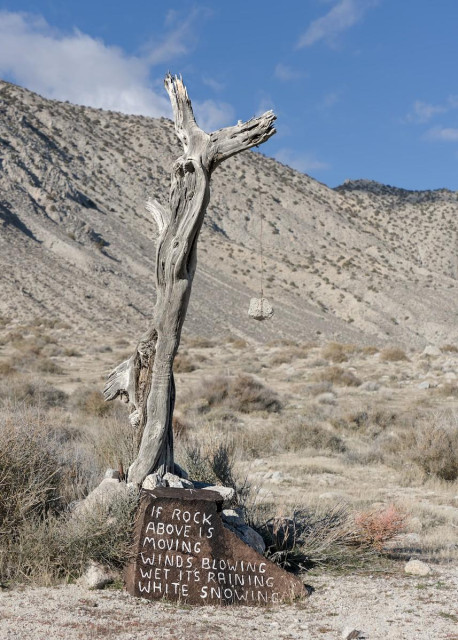 A rock hangs on a thin rope from a very weathered tree branch stuck in the ground. The landscape is light tan-brown winter sagebrush. A mountain is nearby in the background. The sky is blue with some wispy clouds. The brown rock slap at the base of the tree branch with the rock reads, "If rock above is moving winds blowing, wet it's raining, white snowing."