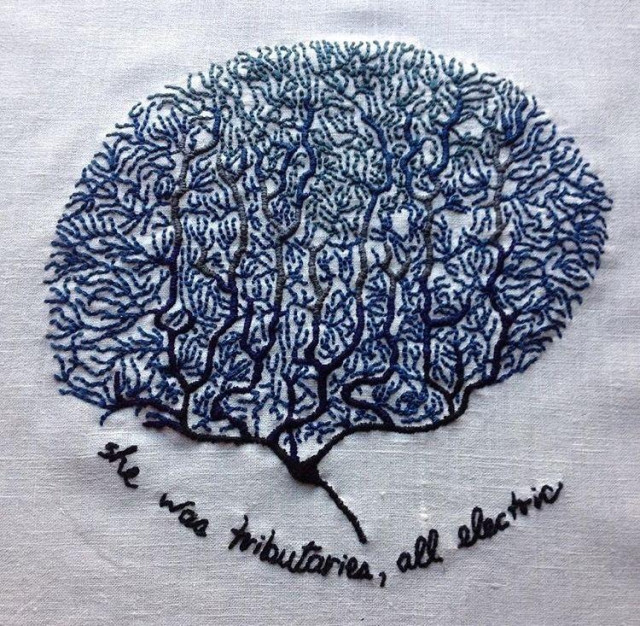 An embroidery on bone white linen. There are many coral-like neurons in the vague shape of a brain embroidered in shades of blue. The bottom section is navy blue and transitions to a lighter blue at the top. The text at the bottom reads: she was tributaries, all electric.