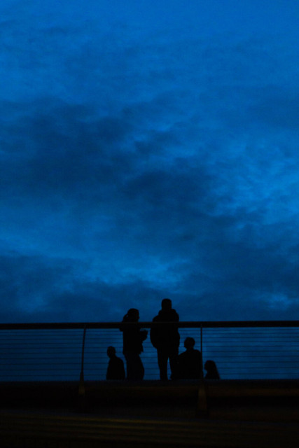 Photo looking up at a deep blue, cloud covered sky. At the bottom is the silhouette of two people standing at a railing, with two others behind them on either side and one more still further back on the right. At the bottom of the picture the underside of the bridge on which they're all standing is very dimly light.