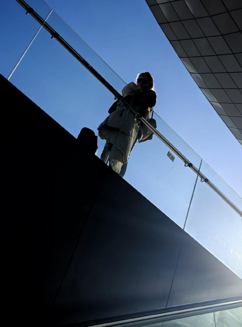 A photo of a person standing on a glass-railed walkway against a clear blue sky, seen from below.