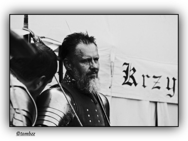 Black and white photo showing two guys dressed as medieval knights, standing in front of a tent marked “KRZYŻACY”, the Teutonic Knights. The main figure is a bearded man in full plate armor: riveted shoulder guards, shiny breastplate, metal collar and gauntlets. His face looks tired and focused, like he’s just come back from a reenacted battle. Sweat and tension give him a raw, authentic presence. The second knight is barely visible, more of a shadow. The soft lighting and subtle vignette create a quiet, serious mood, as if time paused for a moment.