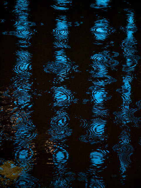 Blue window lights from a tower block hotel reflect in a puddle rippled by heavy rain on a Glasgow street