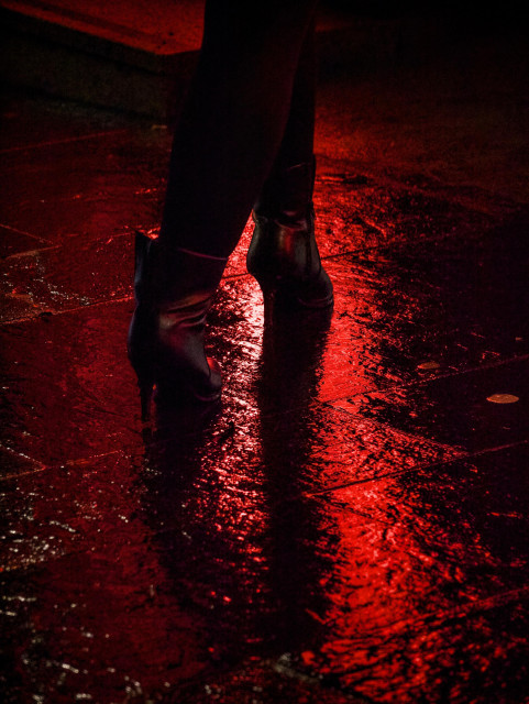 A woman in high-heeled boots walks into a bar where the red neon light is reflected in the soaking wet pavement