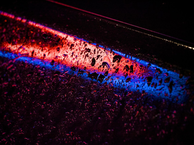 Rain pooling on the curves of a parked car reflects red and blue neon lights from a bar nearby