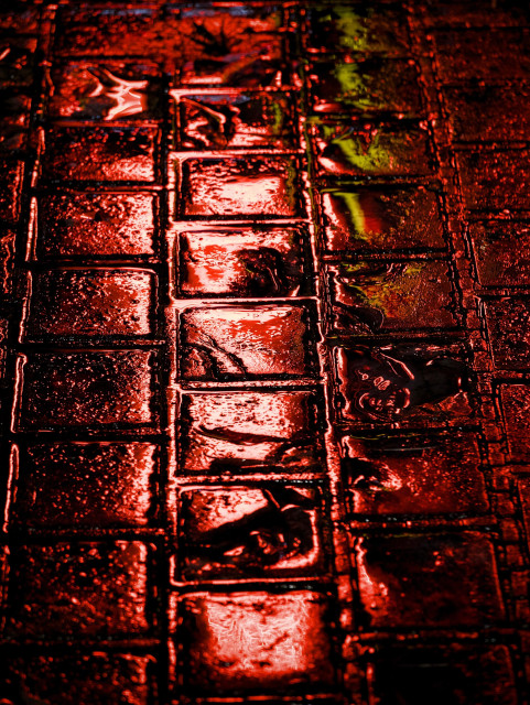 Rainwater pooling on block paving captures and reflects red and yellow neon lights from a takeaway restaurant