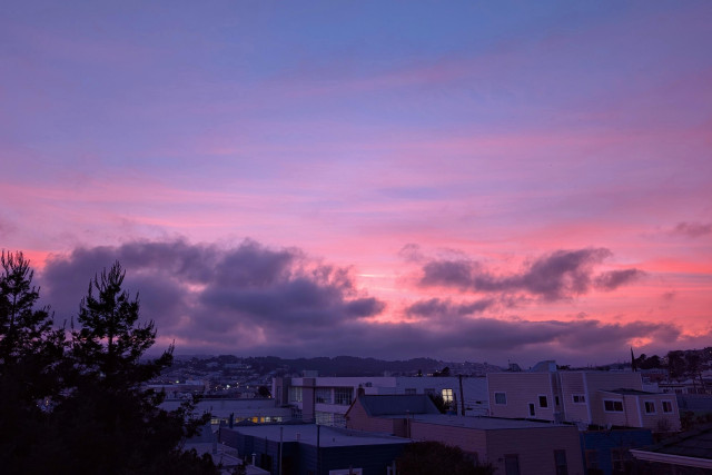 Heavy, amethyst-colored clouds obscure some of the coral and rose-gold colors in the sunset, glimpses of which can be seen beyond the clouds. In the foreground is an urban neighborhood landscape, with silhouetted redwood trees off to the left. CC BY-SA 4.0 Kate Zimmerman.