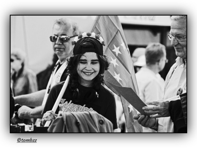 Black-and-white street photo from a pro-EU demonstration. In the center, a young woman with loose hair, wearing a backwards cap decorated with a flower crown. Headphones around her neck, a “Wonder Woman” shirt with the DC logo. She’s smiling and holding a tall EU flag. On the right, an older man with glasses and an EU pin, focused on reading or speaking. Behind her, another man looks toward the camera, curious. The crowd in the background feels dense and diverse, a shared moment of presence and support.