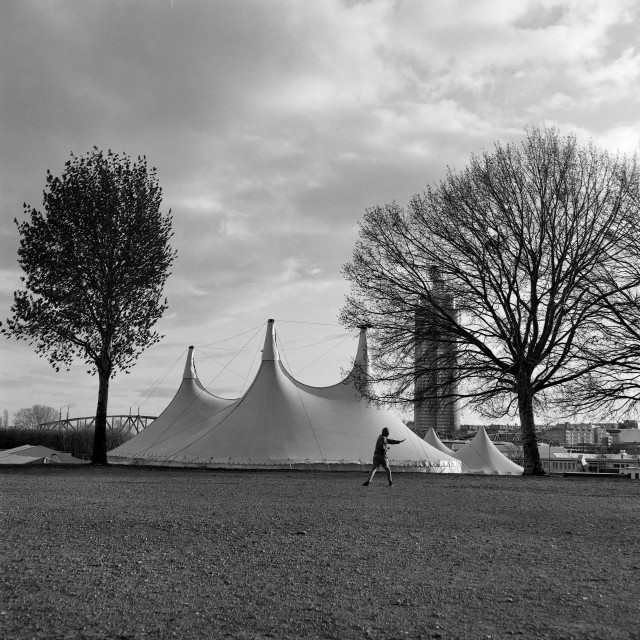The black and white image shows a large tent-like structure with several peaked roofs set in an open area between two tall trees, while a lone person training in the foreground and a cityscape rises in the background under a cloudy sky.