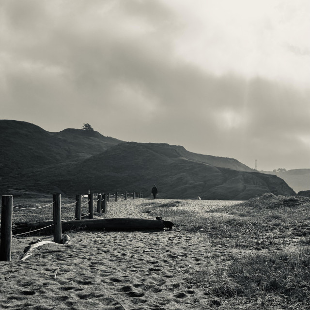 A black and white landscape photograph depicts a dirt path leading towards a cliffside. In the foreground, a weathered wooden fence runs diagonally across the frame, with a large log lying parallel to it. The path is made of dirt and sand, with visible footprints scattered across its surface. Two figures are walking along the path in the distance, approaching the cliff face, which is covered in vegetation and has a gradual slope. The sky is overcast, creating a muted, atmospheric light throughout the scene.