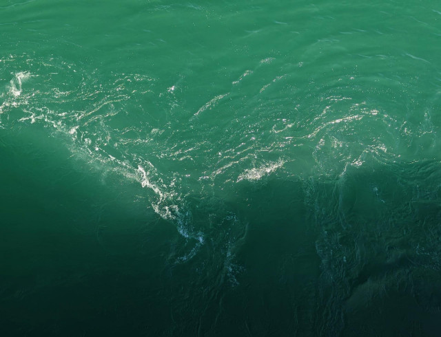 A photo looking down onto green water with visible surface currents and small areas of white foam. Half the photo is in shadow.
