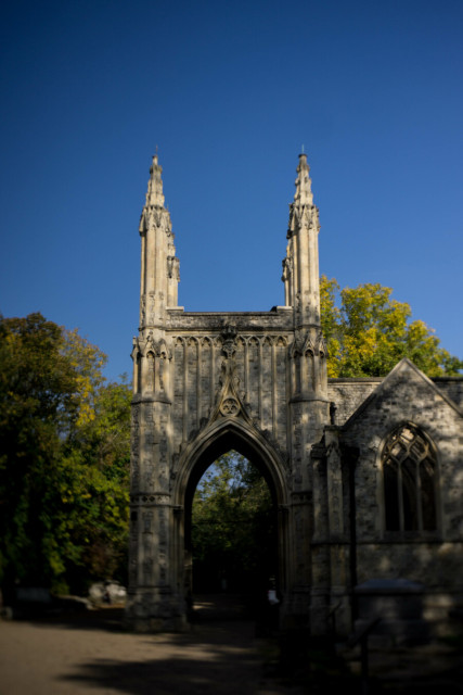 Photo of a short, square Gothic tower in cream-coloured stone, topped with four ornate pinnacles on each corner and large, pointed archways through on each wall at ground level. This is a portico over the entrance to a ruined chapel to the right. The walls are mottled with leaf shadows from the trees that surround the building, which stands in a gravelled open space, the pinnacles alone bathe in pure sunlight. Above is a clear blue sky. The image blurs and distorts the further it gets from the centre.