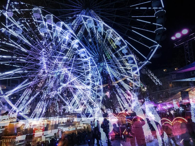 Une photographie nocturne en couleur et en exposition multiple, au format paysage. La scène se passe au pied d'une grande roue illuminée, elle est présente de façon décalée de la gauche vers la droite à trois reprises sur l'image . Au pied du manège, des passants, les chaises d'une terrasse de café . L'ensemble est baigné de lumière artificielle et donne un peu le vertige.