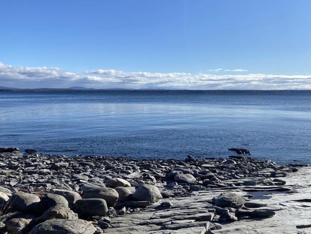 A gray rocky shore in the bottom third of the photo, deep blue lake in the middle third, and blue sky with a few clouds at the horizon in the top third. We're looking East across lake Champlain, standing in New York, looking at Vermont on the far shore. 