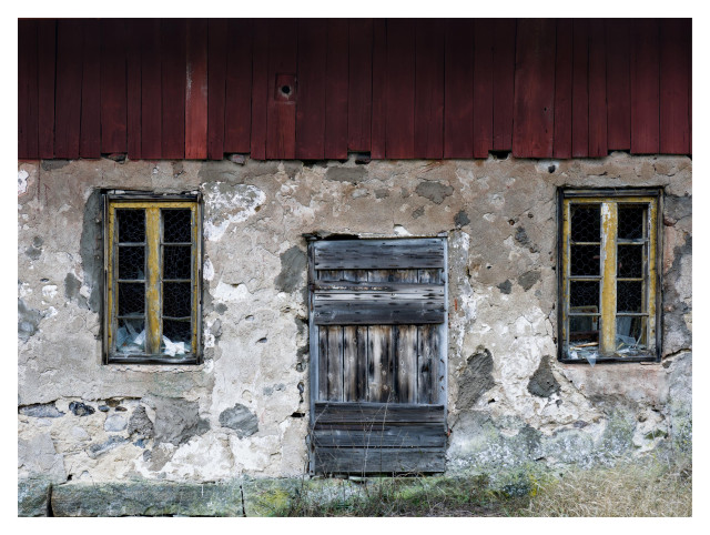 En liggande bild på en sliten ekonomibyggnad med en blek svart trädörr. På båda sidor ett åttadelar fönster med gul träram. Nedre delen av byggnaden i sten med puts, övre i rött trä.