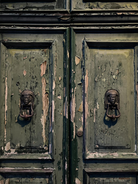Closeup photo of the central panels of a set of panelled wooden double doors. They are painted a dark mossy green, but the paint is largely flaking and fading, revealing flashes of dark blue paint and rough blond wood underneath, and they look like no one has opened them, let alone cared for them, for decades or centuries. There are matching black iron door knockers on both halves of the door, in the shape of stern faces, with the knocker-handle-bits hanging down and casting u-shaped shadows. There is a feeling of dereliction and total neglect, and I think some people might find it a bit spooky-looking and Gothic horror-y, but I think it looks intriguing and mysterious and like an invitation to heart-pounding spectacular adventure, and I would very much like to go inside. 