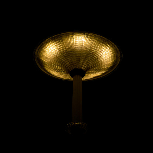 Photograph looking up at a streetlight in the dark. The lamp is a glass disc with ridged segements that curve down to a barely visible post. That light is coming from six bulbs inside the glass can be seen by the regular intervals of brightness towards the edge.