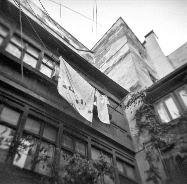 The black-and-white photograph captures an upward view of an old inner courtyard. The camera looks toward the upper floors of weathered buildings with cracked plaster and wooden window frames. Two pieces of laundry—a patterned sheet and a plain cloth—hang loosely from a line stretched between the structures, swaying slightly in the open space above. Vines climb up the walls and around the windows, adding a touch of softness to the otherwise worn architecture. The edges of the image fall into a gentle blur, giving the scene a nostalgic, almost dreamlike atmosphere