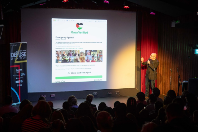 Me (white passing guy with short hair wearing a brown tweed jacket, black t-shirt, and jeans), presenting to a small audience in front of a screen that has the Gaza Verified web site on it.