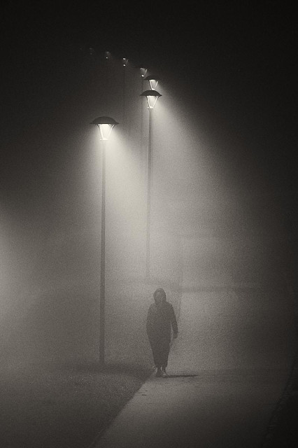 A lone figure in a dark hooded coat walks towards the camera along a narrow path at night, almost swallowed by the dense fog that softens every edge. Tall streetlamps stand in a diagonal line receding into the distance from the top left of the frame, each casting a narrow beam of warm light that pierces the mist and fades into the darkness. The person's face is hidden in shadow, their body only partially defined, adding to the sense of anonymity and unease. The grainy texture and monochrome tones make the scene feel both cinematic and isolated.