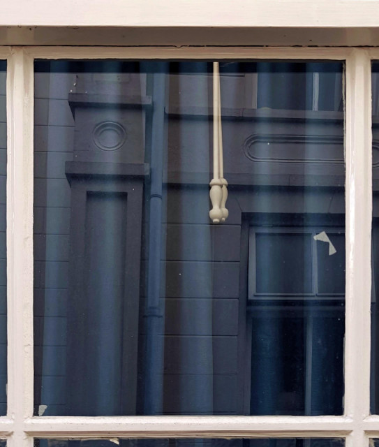 A photo showing a part of a window from the outside with white wooden frames and closed blue curtains. The reflection in the glasses shows a building facade across the street.