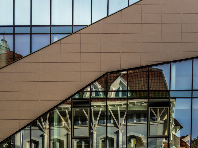 In stark verspiegelter Fassade (die auch einen geschwungen geschlossenen Bereich zeigt) spiegelt sich ein gegenüber liegendes Gebäude mit Fenstern. 