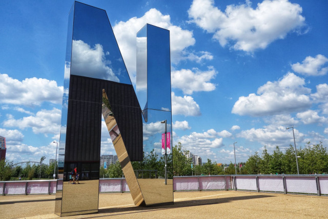 This image showcases a large, reflective metallic sculpture of the letter "N" from the "R U N" installation located in Queen Elizabeth Olympic Park, London. The sculpture stands prominently against a bright blue sky dotted with fluffy white clouds. The polished surface of the letter reflects the sky, clouds, part of the Copper Box Arena, a street lamp, people walking by, and surrounding greenery, creating a dynamic visual effect. The ground is covered in gravel, and the installation is enclosed by a low fence. In the background, trees and urban structures are visible, adding context to the park setting.