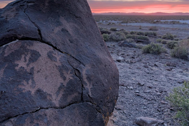 A color landscape photo of a petroglyph and a desert sunset. A large frame filling boulder occupies the left side of the frame. On the boulder are several petroglyphs. The largest one appears to be a creature with four legs and head and a tail shaped somewhat like an arrow. The other smaller glyphs are simple geometric shapes. The right side of the frame is a desert landscape of sagebrush extending out to a far distant range of mountains on the horizon. The thin strip of sky visible is red and orange with late sunset colors.