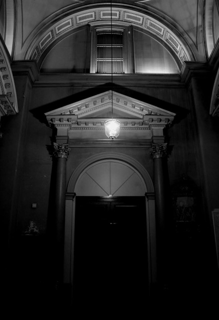 A black and white photograph of a monumental doorway inside a church, in a dark corner illuminated only by a lamp suspended directly above. The double door is surmounted by an arch and framed by Corinthian columns with a triangular pediment. Above the doorway is a rectangular window set within a decorated arch.

Photographie noir et black d'une porte monumentale à l'intérieur d'une église, dans un coin sombre et éclairé seulement par une lampe suspendue juste au-dessus. La double porte est surmontée d'une arche et encadrée de colonnes corinthiennes avec un fronton triangulaire. En haut de la porte se trouve une fenêtre rectangulaire au creux d'une arche décorée.