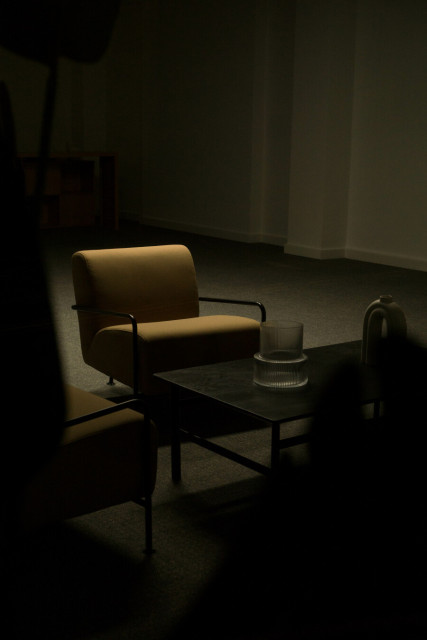 Photo of a shadowy room with light falling on a low chair in front of a dark coffee table. On the table is what looks to be a large glass candle holder, empty. Dark indistinct shapes close to the camera obscure the image along the lower edge.