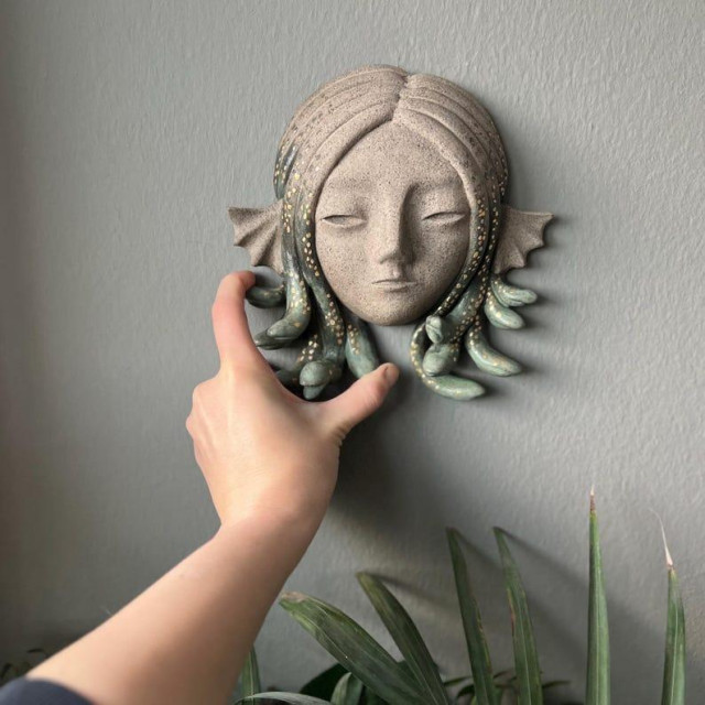 An mostly unglazed clay mask hanging on a wall. It depicts a woman's head with some fin like ears and snakes as hair. The heads of the snakes are glazed green and decorated with gold.