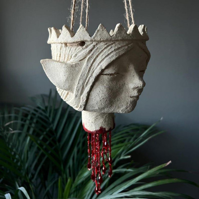 And mostly unglazed clay pot hanging on some threads. The surface is ruff, reminding of sandstone. It depicts a elf's head, wearing a crown. The neck is severed and red beads,  looking like blood, are hanging from it.