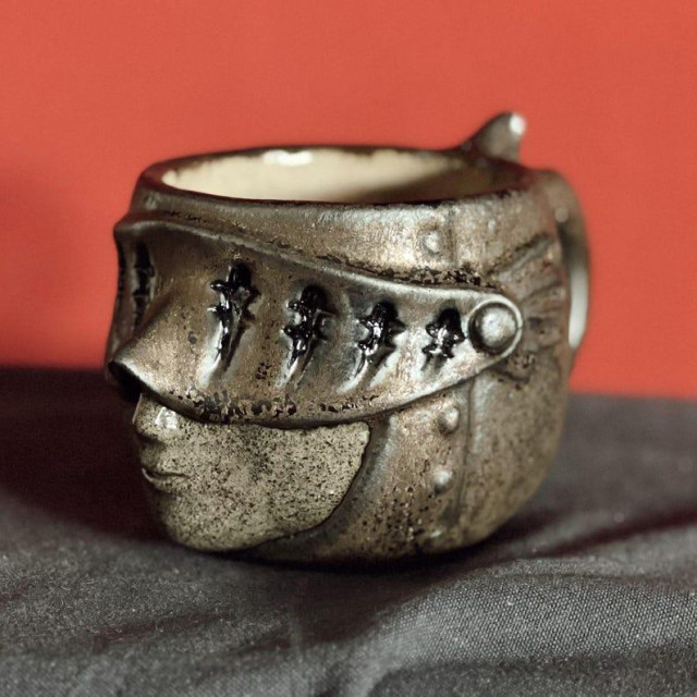 A cup in the shape of a head in a medieval helmet. The glaze looks metallic.