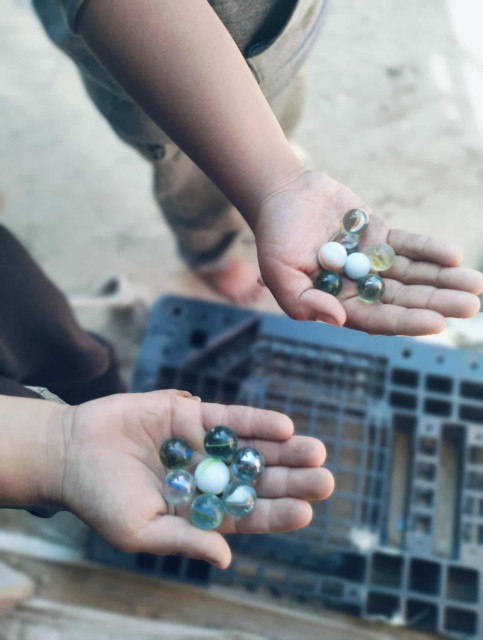 Two young children holding multiple colorful marbles in open hands, with one child's arm and legs visible in the background and a blurry object in the foreground.