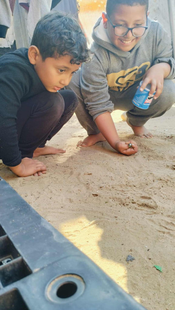 Two young boys squatting on sand, Saji is looking intently at a marble held by his brother Elias, who is smiling and holding a Pringles can.