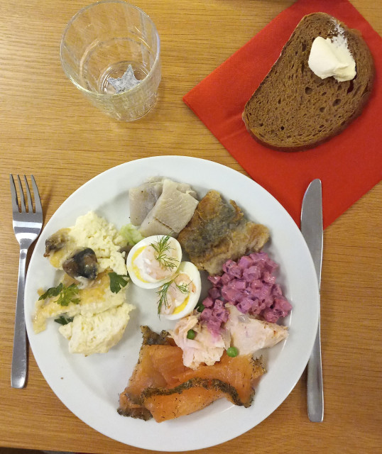 En vörtskiva med smör på en röd servett. En tallrik med lax, omelett, ägghalva,  rödbetssallad och sill