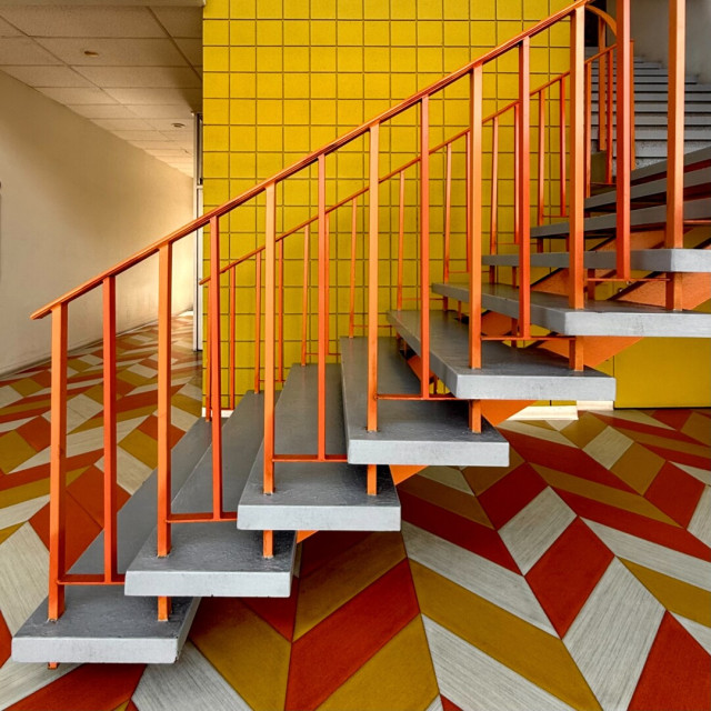 Interior stairwell of a commercial building with orange railings, a yellow tile wall in the background, and orange, yellow, and white tile floors in a geometric pattern.