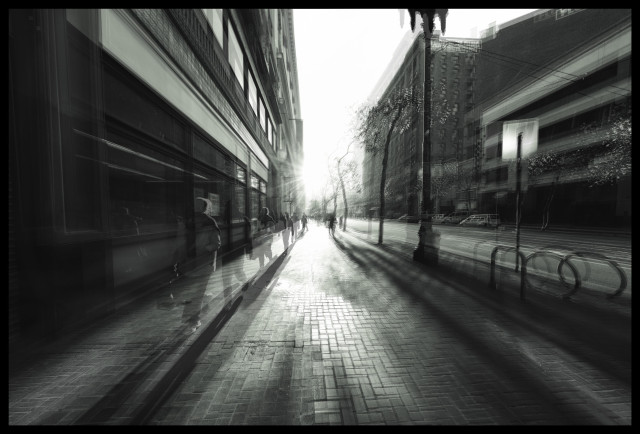 A monochrome triple-exposure photograph of a brick sidewalk and city street on a late fall day. Taken downtown in San Francisco, on Market Street near Powell, there are dark, mid-rise buildings on either side of the photo. The sun is low on the horizon, shining directly into the camera... creating glare while casting very long shadows as the ghosts of people walk towards the camera. The result is jittery, moody, and very urban.