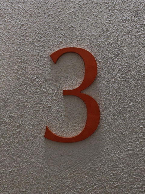 A photo of a red/orange number 3 mounted on a textured light-colored wall.