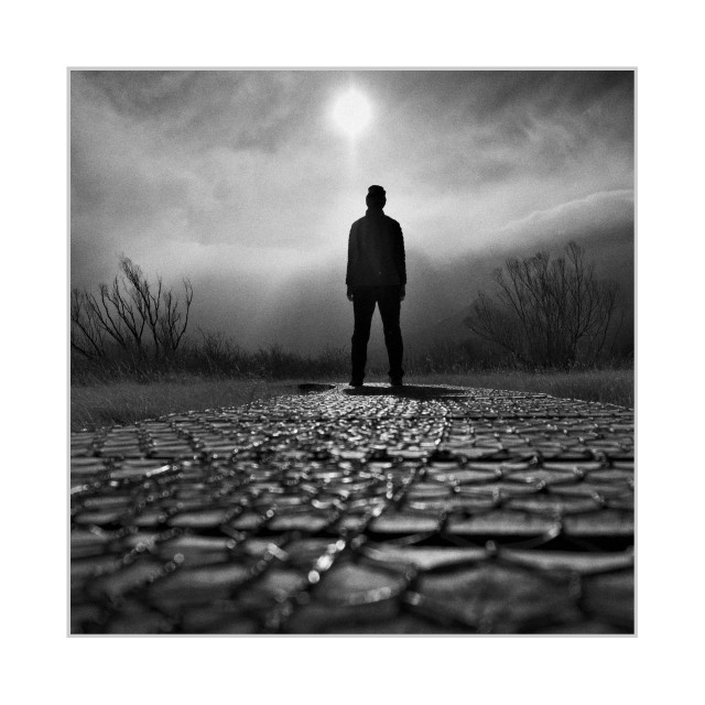 Square black-and-white photograph. In the foreground, a path made of wooden boards covered with wire mesh stretches toward the background. The mesh catches the light and creates a pattern of small, bright highlights. At the far end of the path stands a single person in silhouette, facing away from the camera. Above them, the sun shines through mist or low clouds, forming a vertical beam of light. Sparse, leafless shrubs frame the scene on both sides, while fog softens the distant landscape. The low viewpoint emphasizes the textured surface leading directly to the figure and the bright sun above.