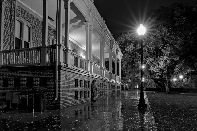 A black and white landscape photo of an empty urban scene at night. It has been raining and everything is wet and reflective. A wide sidewalk extends out from the bottom left to about center frame. A large Victorian style building is on the left but all we see is the brickwork with numerous small windows and a large porch about six feet off of the ground. The porch is bordered by a white wood railing. Old style street lamps on posts border the sidewalk on the right. grass and trees dominate the right side of the frame. A man mysteriously stands against the brick part of the building looking out toward the grass area. He is far away, but we can see he wears a flop hat and a raincoat.