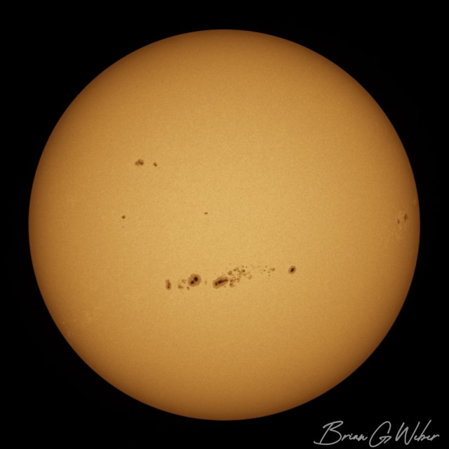 A full disk monochrome image of the sun, stacked from 750 frames of video and colorized orange. The main feature is a tight band of sunspots right in the middle just below the equator.